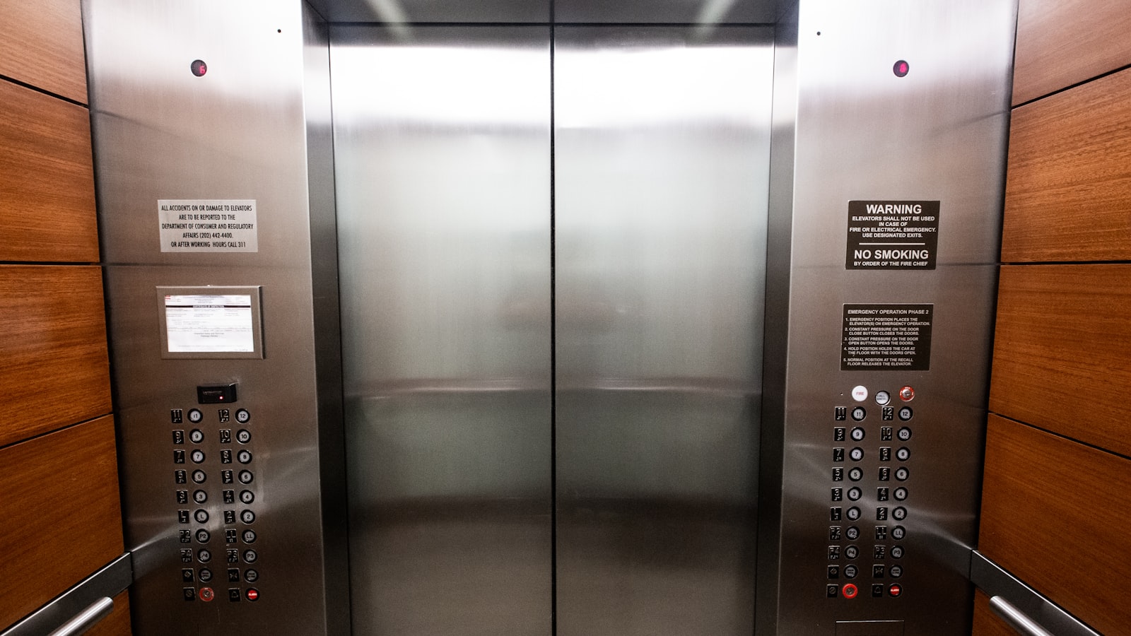 Stainless steel elevator doors with floor buttons and a framed inspection notice on the panel