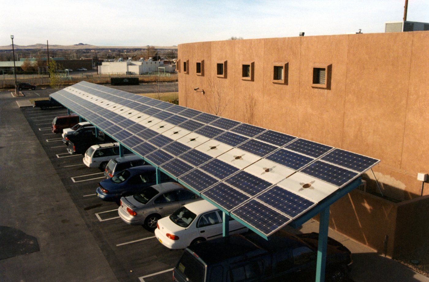 A row of solar panels mounted as a carport canopy shades parked cars beside a low adobe-coloured building.