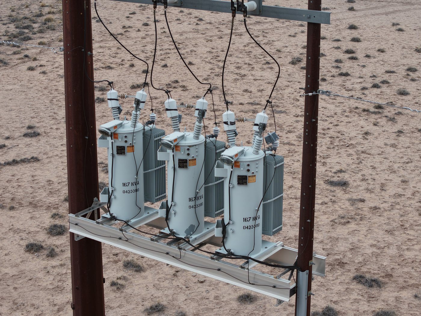 Three pole-mounted electrical transformers hang from a steel crossarm against a pale sky, insulators and drop wires visible.