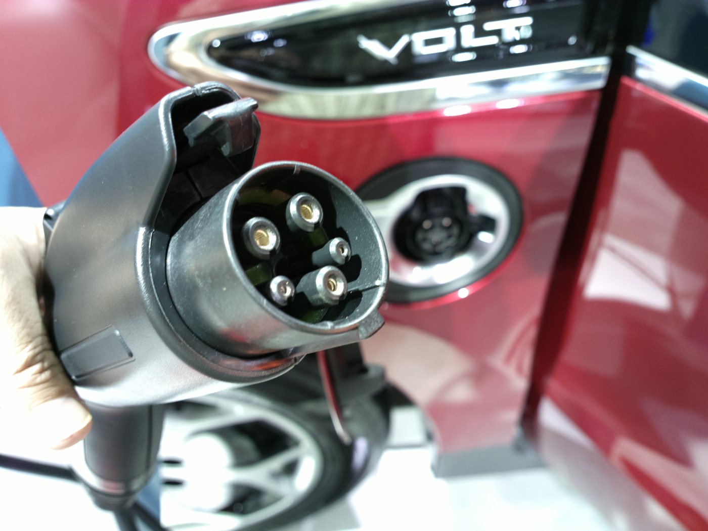 A close view of an electric vehicle charging connector held beside the open charge port of a red car.