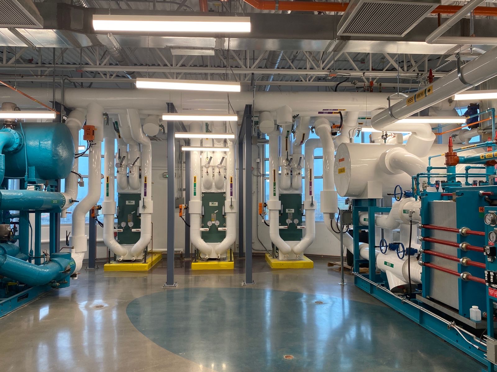 A central plant room under fluorescent light, with plate heat exchangers on yellow housekeeping pads and insulated white piping running to teal chiller skids.