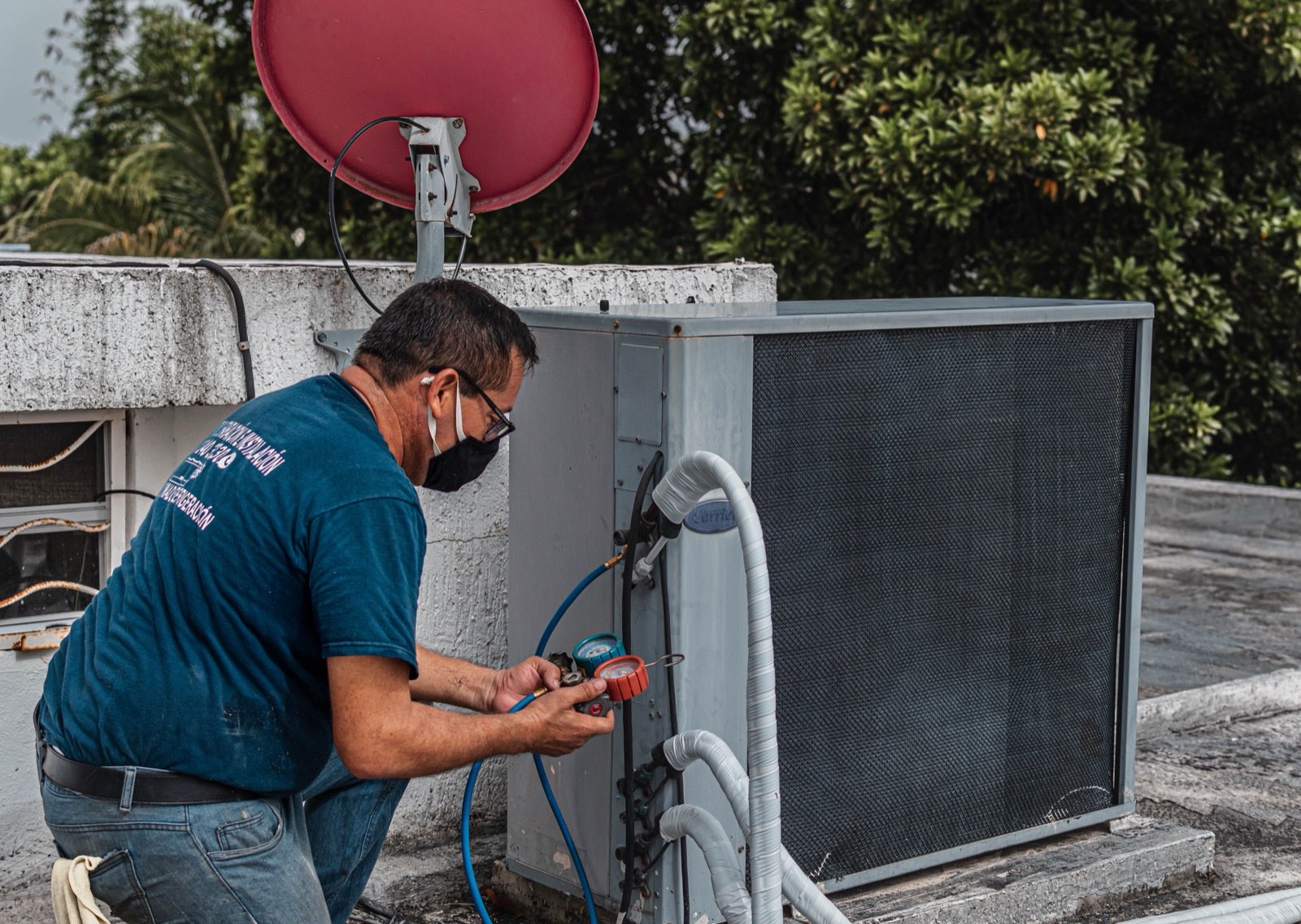 A masked technician kneels on a rooftop reading a manifold gauge set hooked to a gray Carrier condensing unit, hoses running to the service valves.