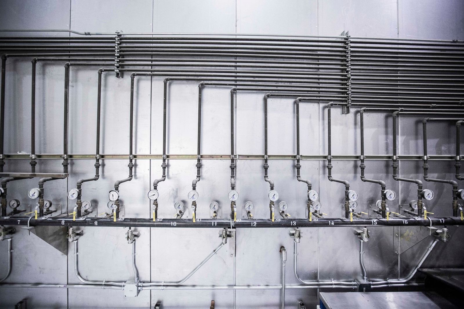 A wall of numbered pipe risers off a black header main, each drop carrying a pressure gauge and a yellow-handled valve, inside an industrial plant.