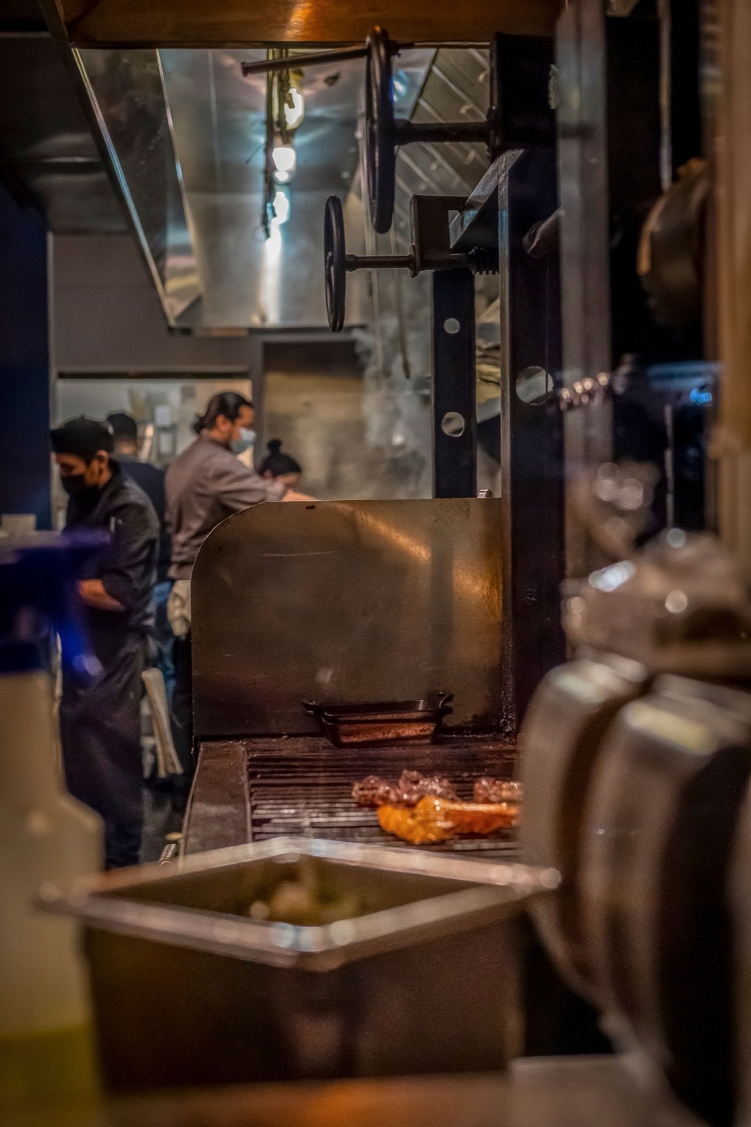 Cooks work a grill line under a stainless exhaust hood as smoke rises off the meat into the canopy.