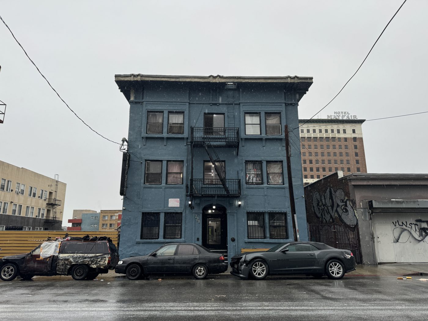A three-storey painted brick apartment building with a fire escape on its face, cars parked along the wet street in front of it.