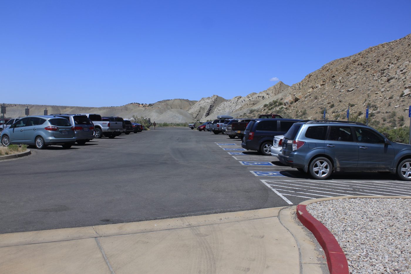 An asphalt parking lot with rows of parked cars, a painted accessible stall and red-painted curb in the foreground and dry hills behind.