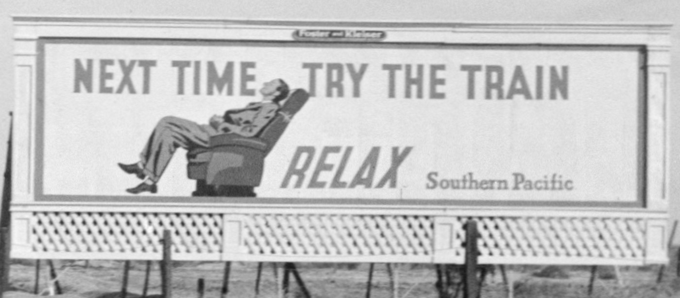 A black-and-white photograph of a painted roadside billboard showing a man reclining in an armchair beside the words next time try the train.